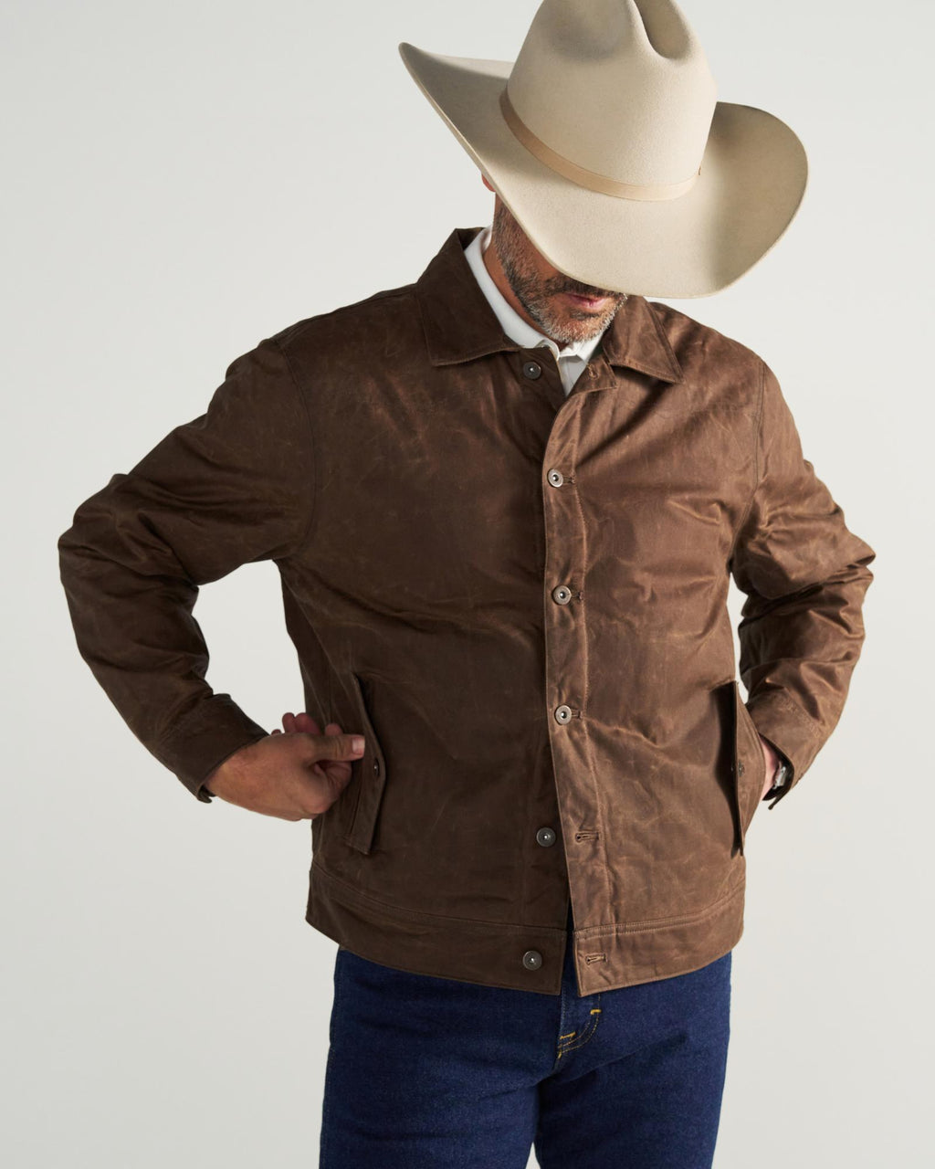 A person wearing a beige cowboy hat, brown button-up jacket, and blue jeans stands with hands in pockets against a plain background.
