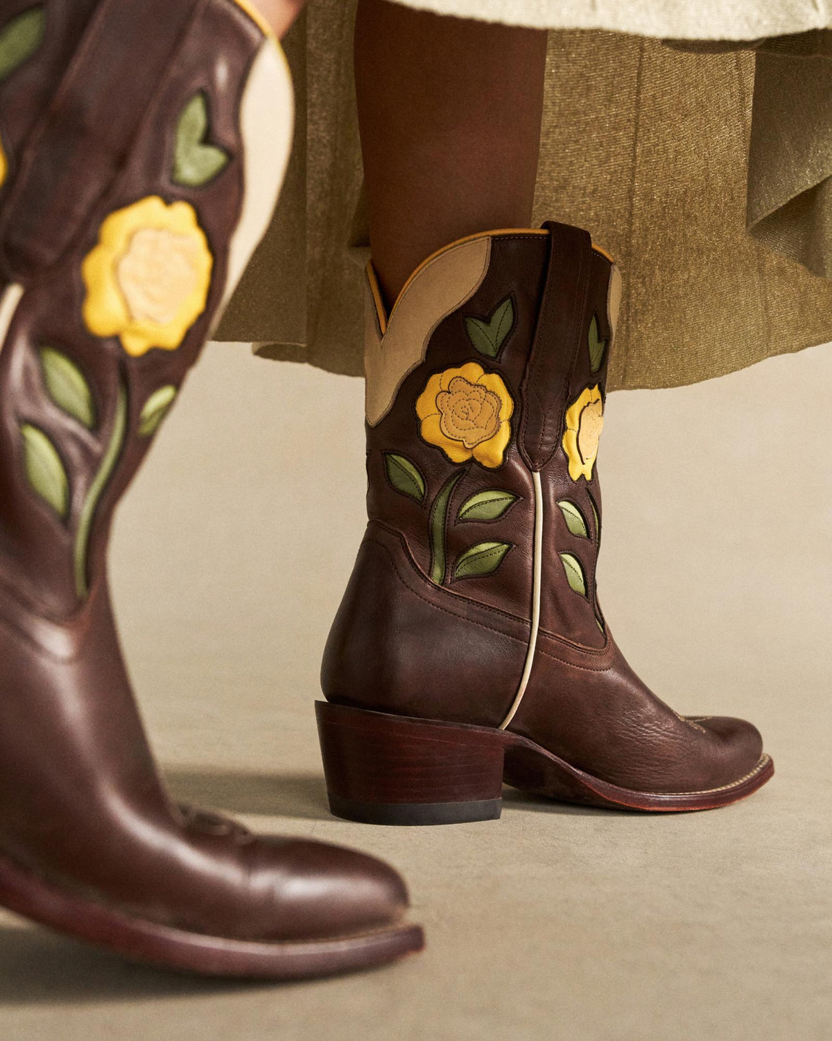The Jolene in Truffle Cowhide. Dark brown truffle cowhide cowboy boots with yellow rose and green leaf appliqué, worn with an olive skirt. A taller matching boot is visible in the foreground.