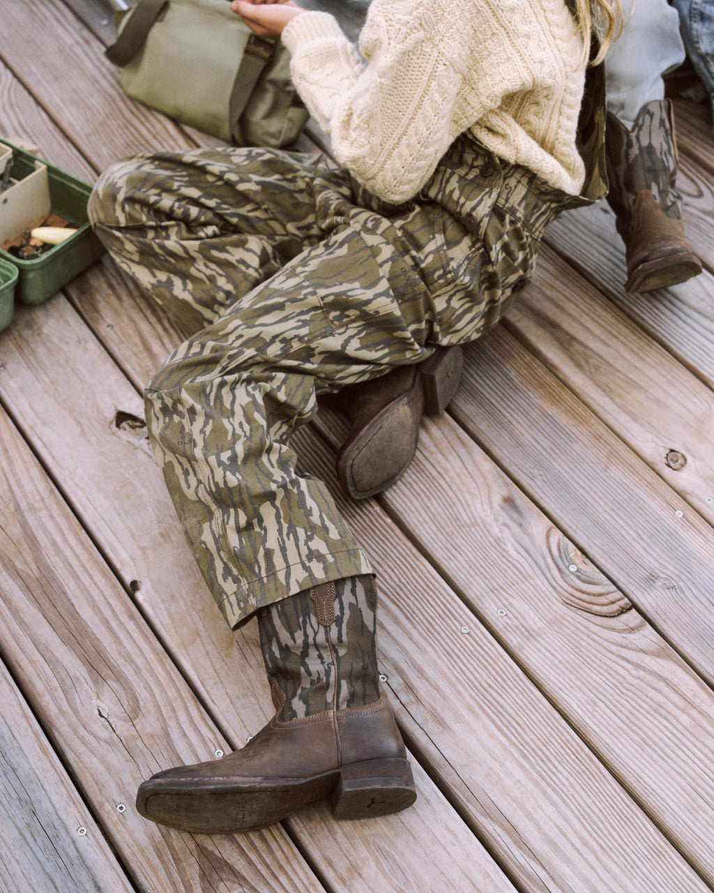 Person wearing camouflage pants, brown boots, and a cream sweater sitting on a wooden deck next to a green bag and small open box.