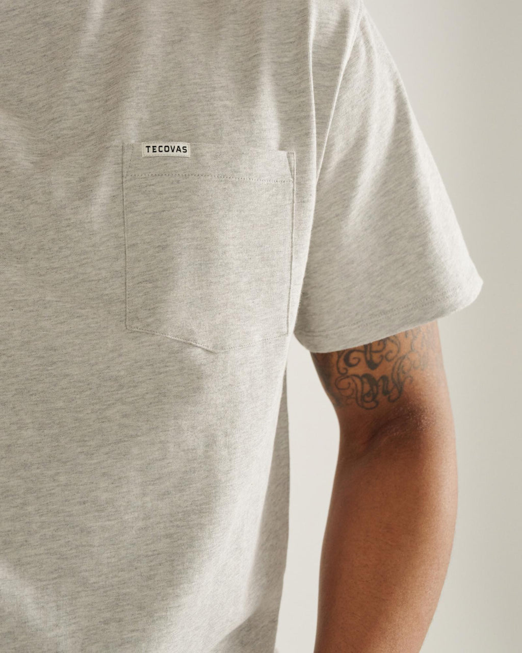 A person wearing a light grey pocket t-shirt with "Tecovas" on the pocket, showing part of their arm with a tattoo.
