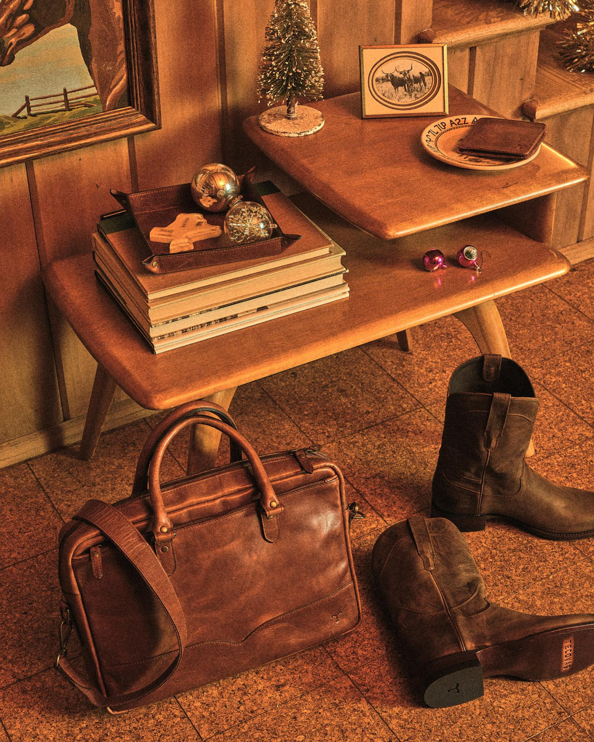 A wooden side table with books, ornaments, and decor sits in a room with a framed painting, leather bag, and brown boots on the tiled floor.