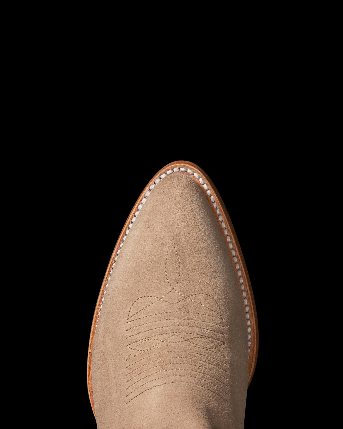 Close-up of a tan suede cowboy boot on a black background, featuring detailed stitching on the upper part and white trim along the edge.