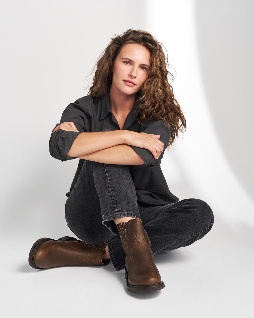 The Chelsea in Cafe Cowhide. A woman with wavy brown hair sits on the floor with her arms crossed, wearing a dark button-down shirt, dark jeans, and brown leather boots against a white studio background—her look is effortlessly stylish, reminiscent of The Chelsea Cowhide collection.