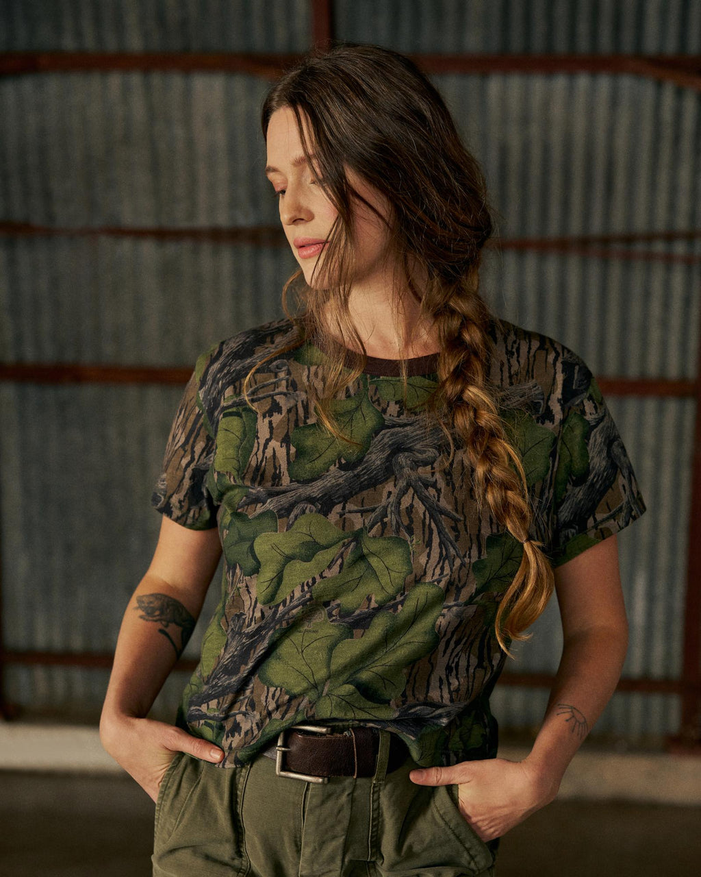 A woman with a long braid, wearing a camouflage t-shirt and green pants, stands indoors with her hands in her pockets, looking down.
