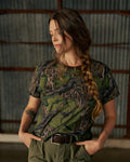 A woman with a long braid, wearing a camouflage t-shirt and green pants, stands indoors with her hands in her pockets, looking down.