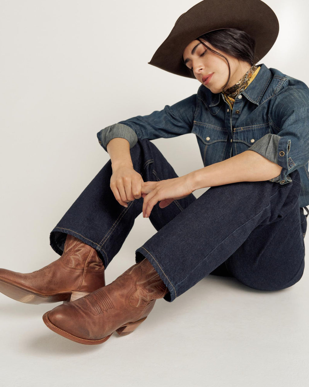 Person wearing a cowboy hat, denim shirt, jeans, and brown cowboy boots sits on the floor with eyes closed and head tilted down.