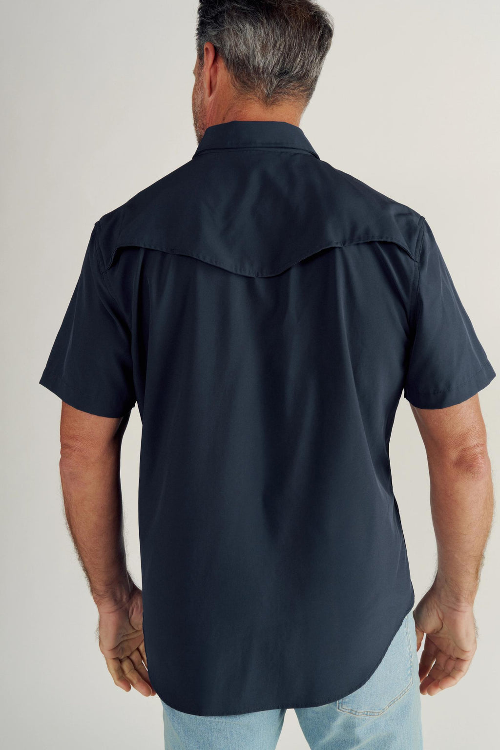Man wearing a short-sleeve, dark button-up shirt and light blue jeans, shown from the back against a plain background.