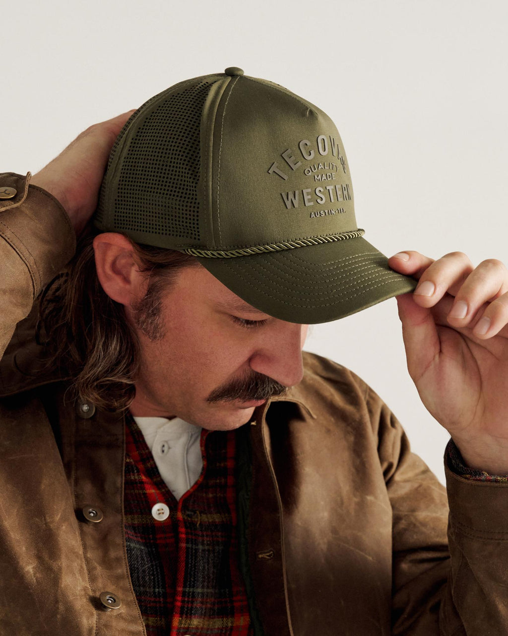 A man wearing a green trucker hat, brown jacket, and plaid shirt looks downward while adjusting the brim of his hat with one hand.