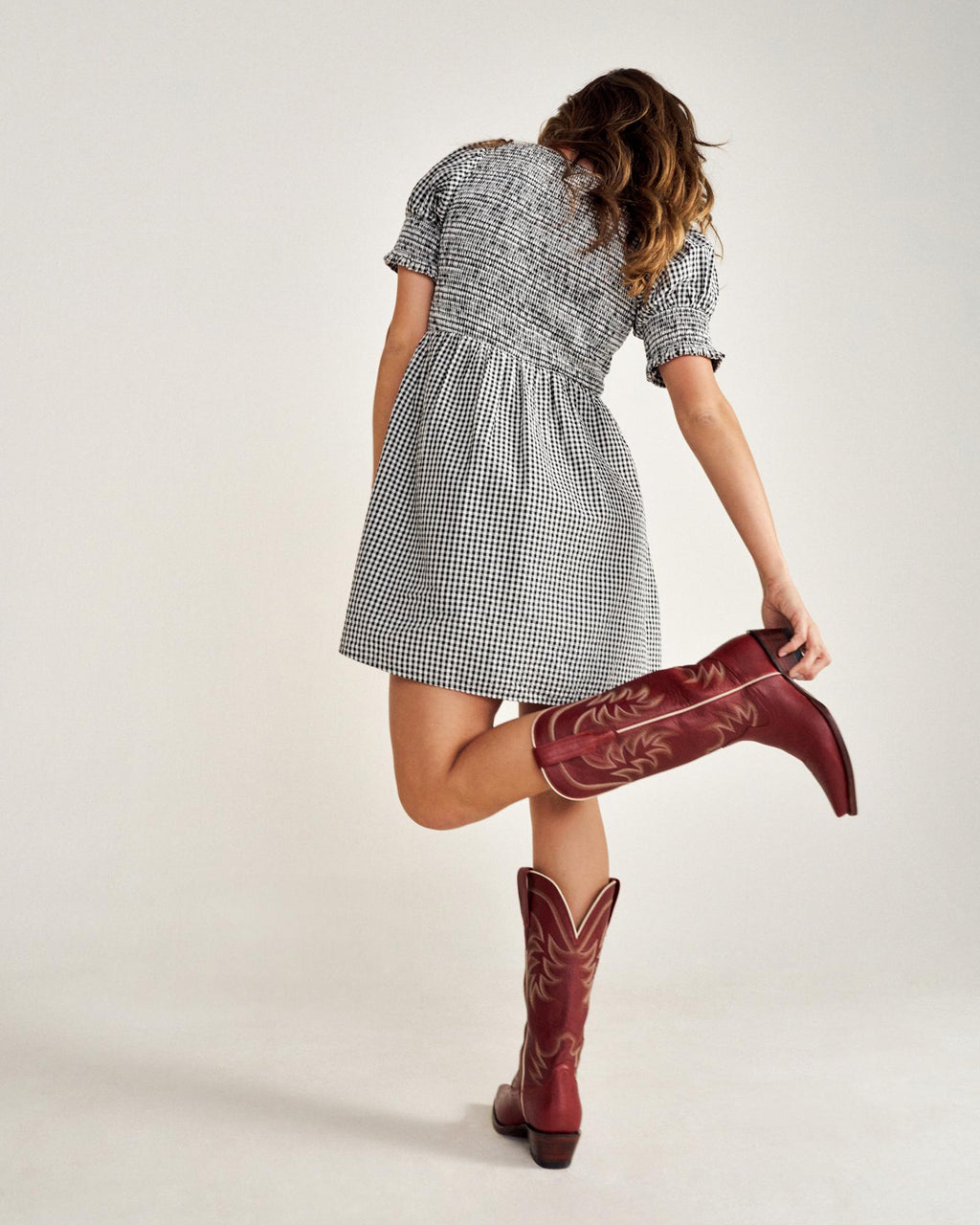 A person in a checkered dress and red cowboy boots poses with one foot lifted, facing away from the camera.