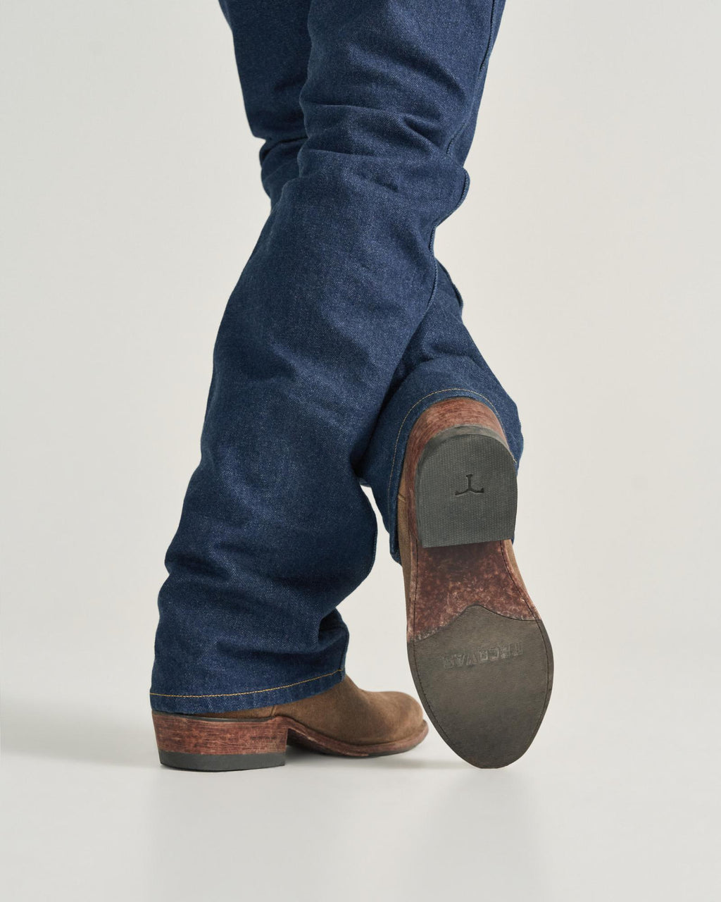The Johnny in Gravel Suede. Person wearing blue jeans and brown Gravel Suede cowboy boots, shown from behind with one leg crossed over the other, displaying the sole of one boot.