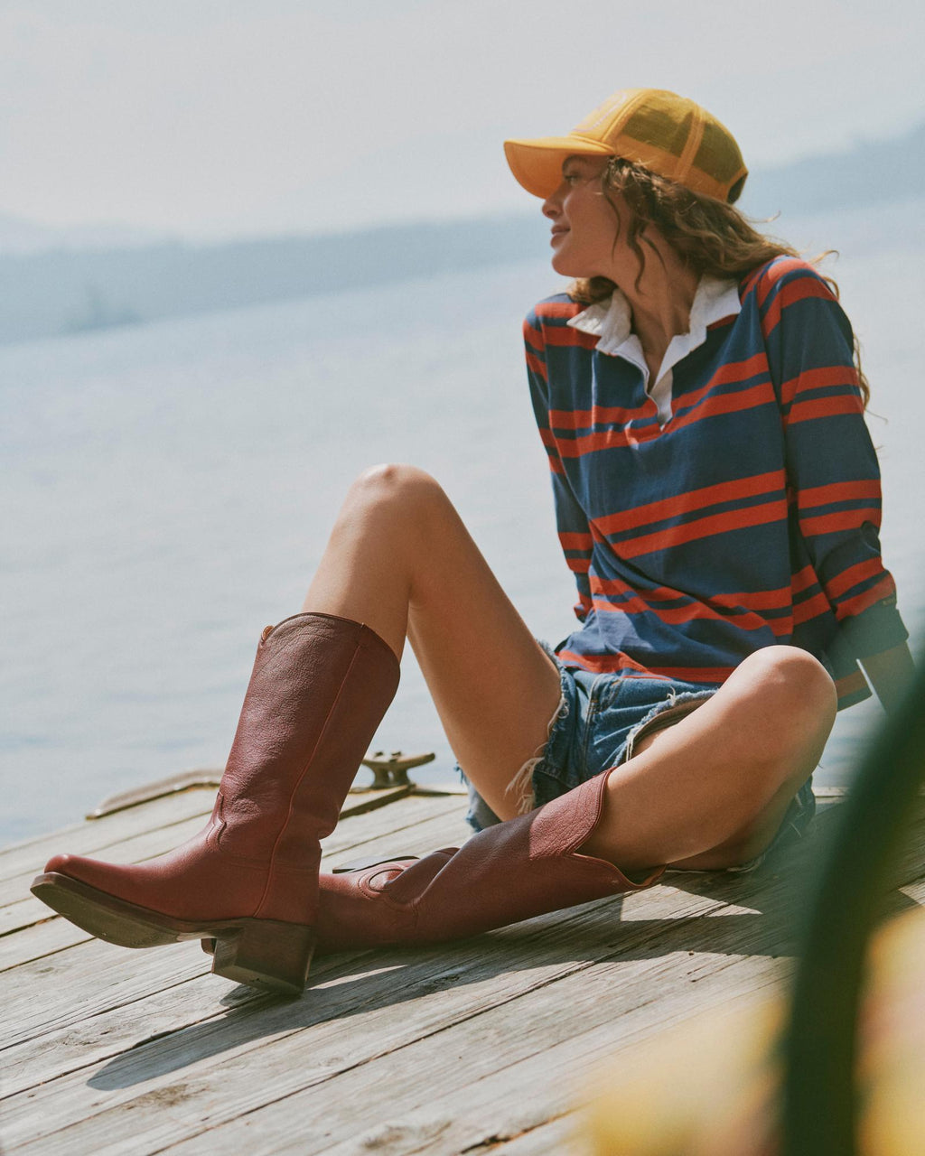The Charlie in Scarlet Cowhide. Person sitting on a wooden dock by the water, wearing a striped shirt, yellow cap, denim shorts, and tall scarlet cowhide boots, looking to the side.