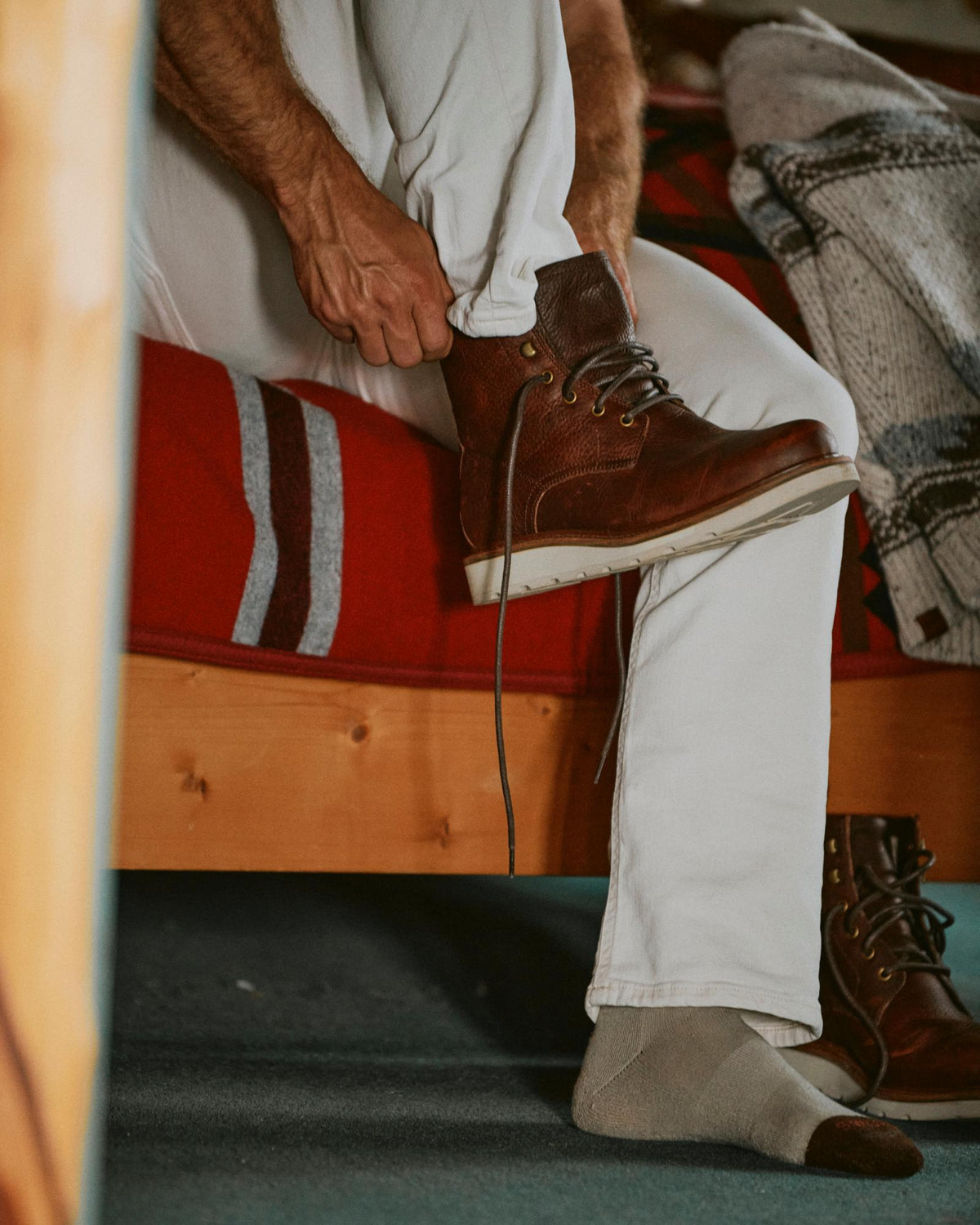 The Knox Lace Up in Cognac Cowhide. A person wearing white pants putting on a cognac Knox lace-up cowhide boot.
