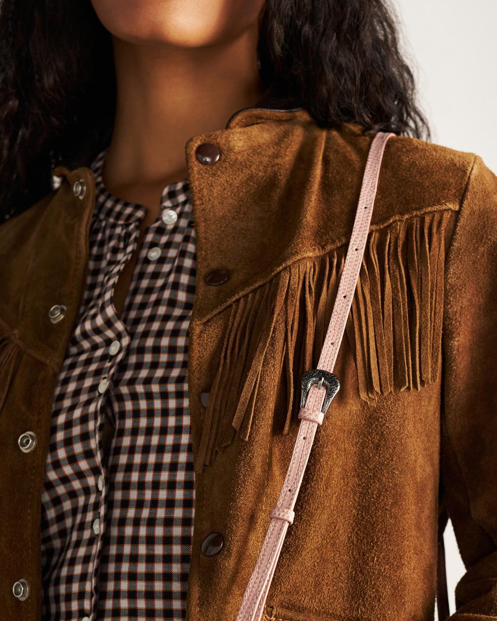 A person wearing a brown suede fringed jacket over a checked shirt with a pink strap across the shoulder.