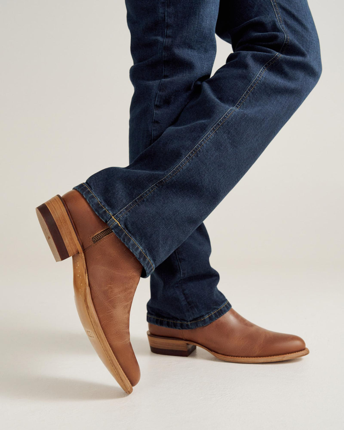 A person wearing blue jeans and brown leather boots with stacked heels stands with one leg crossed over the other against a plain background.