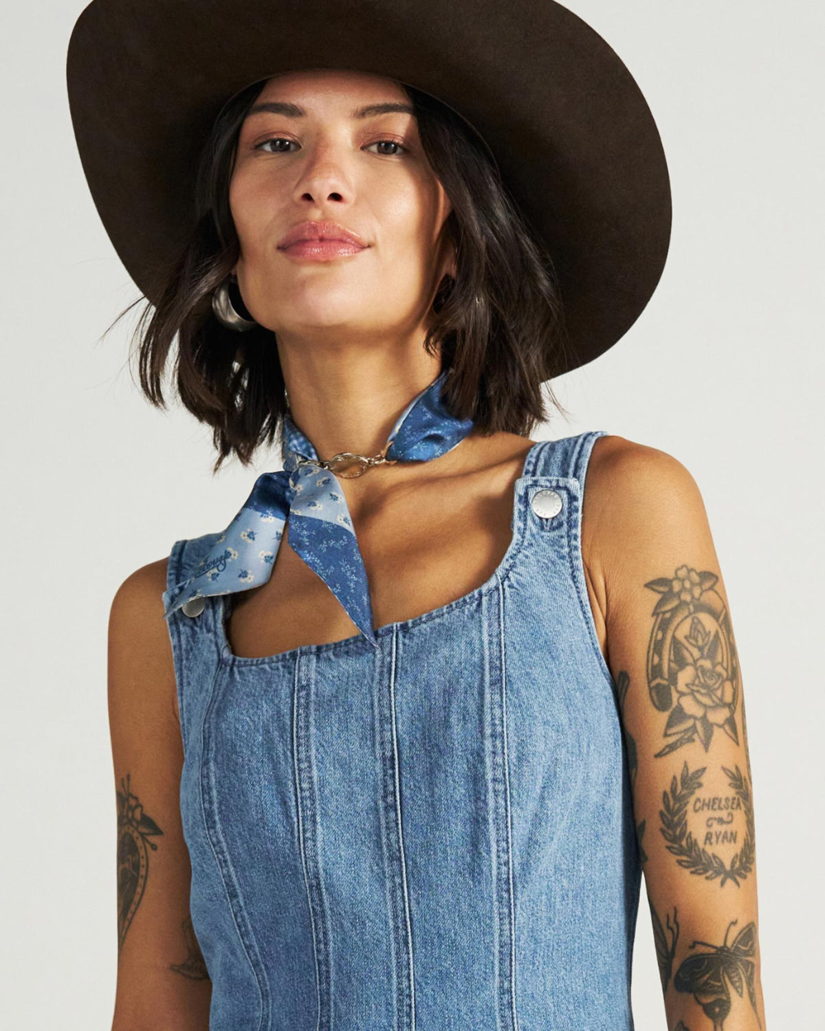 A woman with tattoos wears a sleeveless denim dress, a blue patterned neck scarf, and a wide-brimmed hat, looking confidently at the camera.