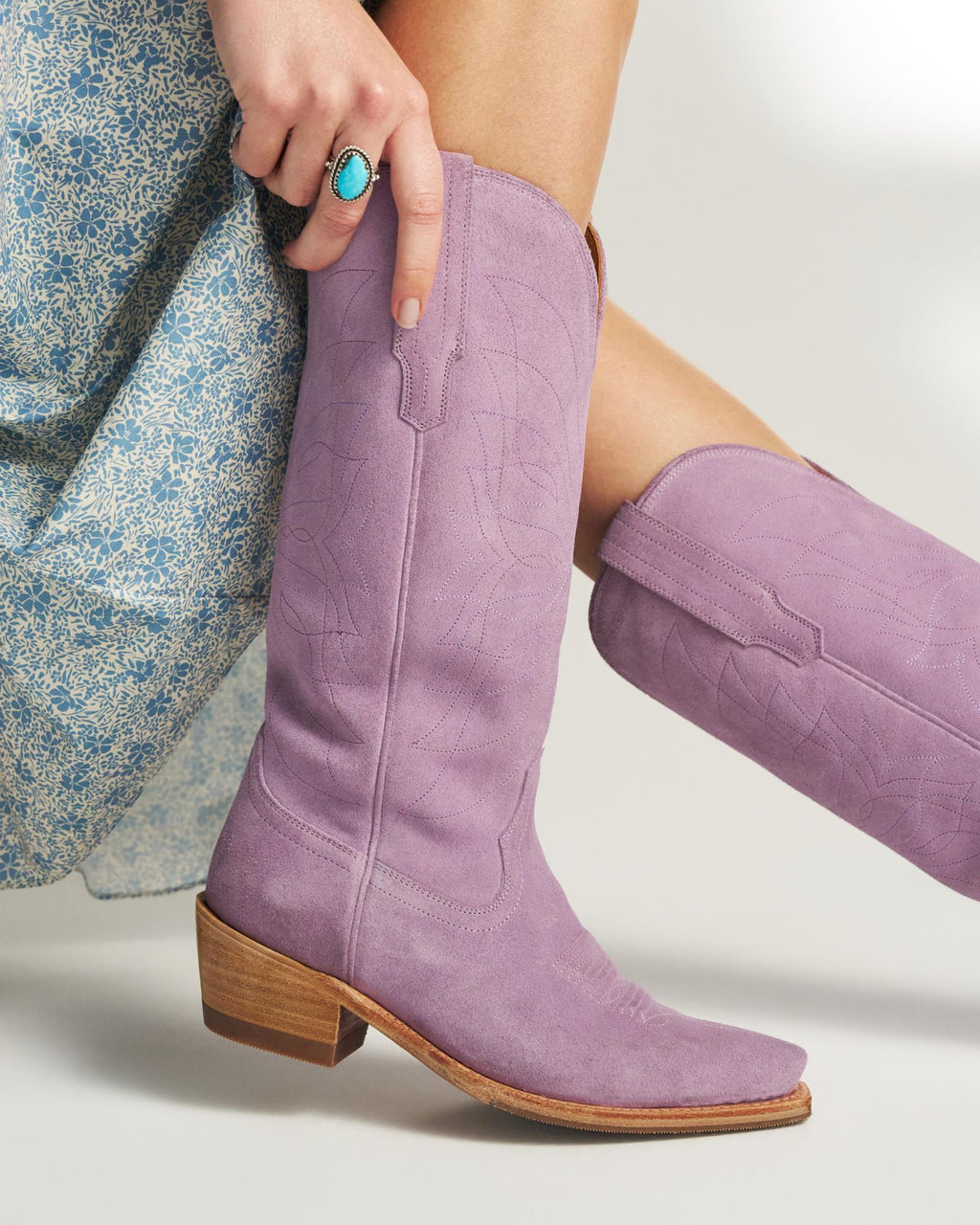 The Annie in Lavender Suede. A person wearing a blue patterned dress and a turquoise ring adjusts The Annie Suede mid-calf, light purple cowboy boots with wooden heels.