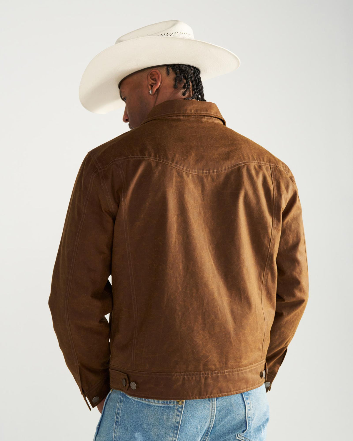 A person seen from behind wearing a white cowboy hat and a brown jacket over blue jeans.