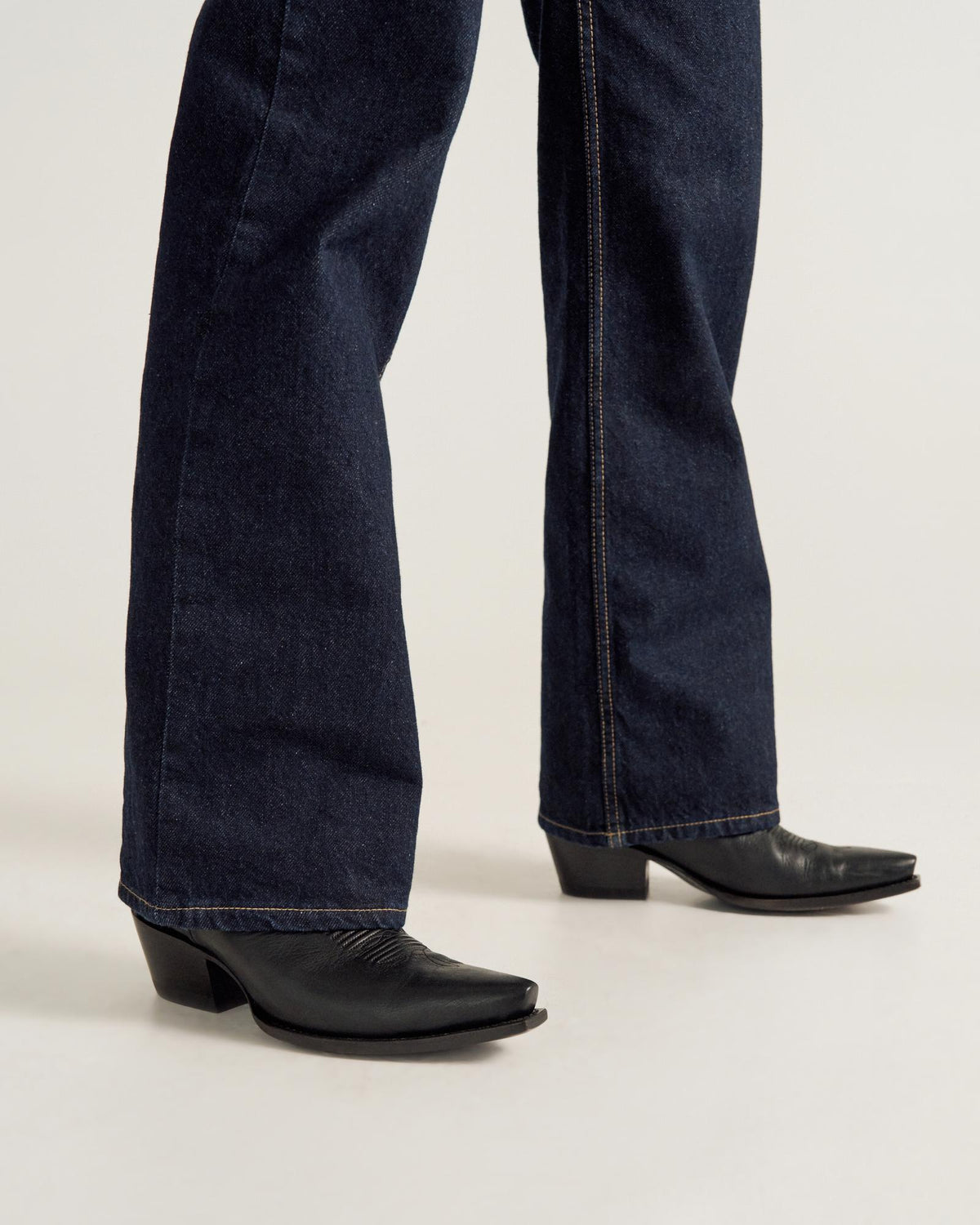 A person wearing dark blue bootcut jeans and black heeled boots stands on a light-colored floor.