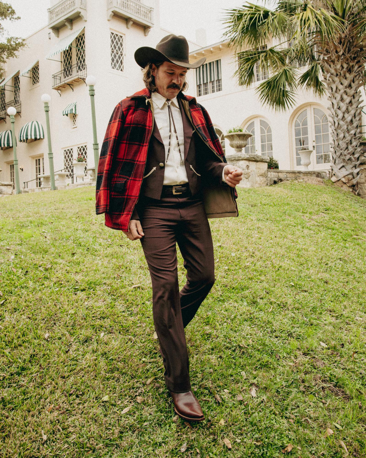 Man in cowboy hat and red plaid jacket walks on grass in front of a large building with arched windows and palm trees.