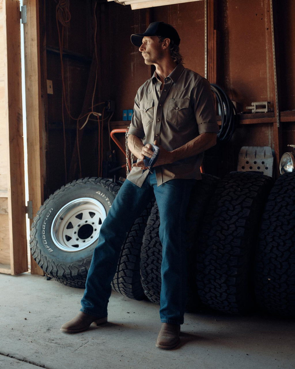 A man in a cap, button-up shirt, jeans, and boots stands in a garage next to four stacked tires, looking toward a source of light.