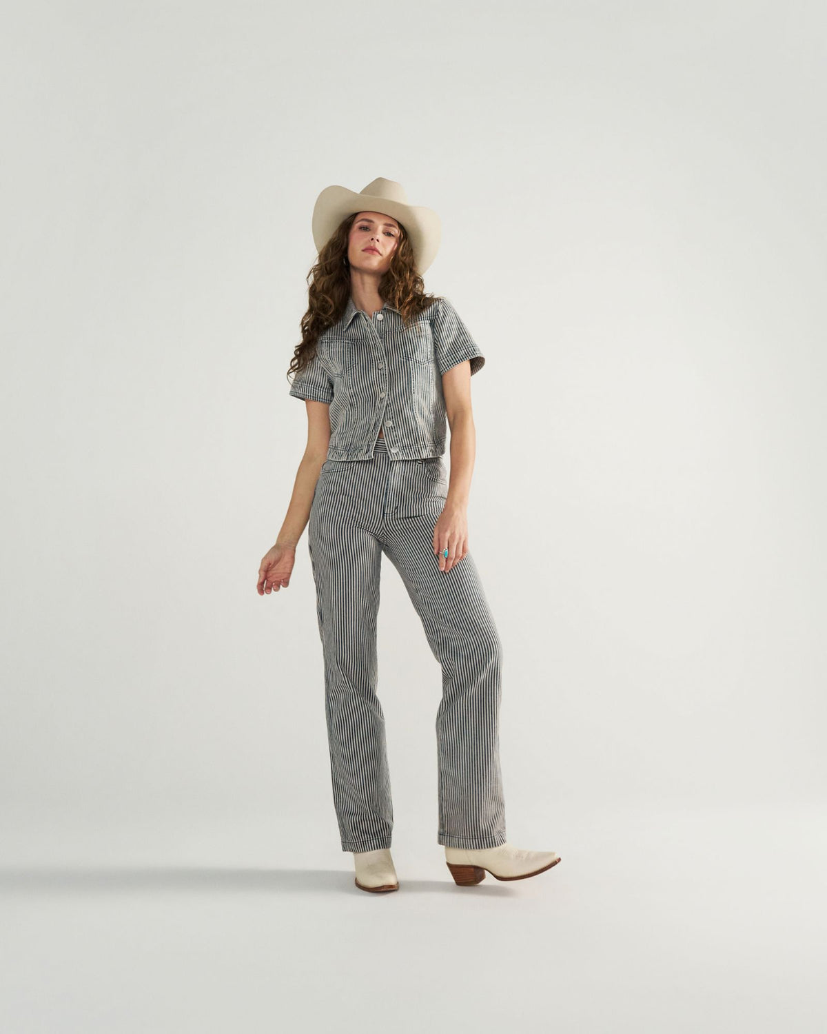 A woman wearing a white cowboy hat, short-sleeve striped button-up shirt, matching striped pants, and white boots stands against a plain white background.