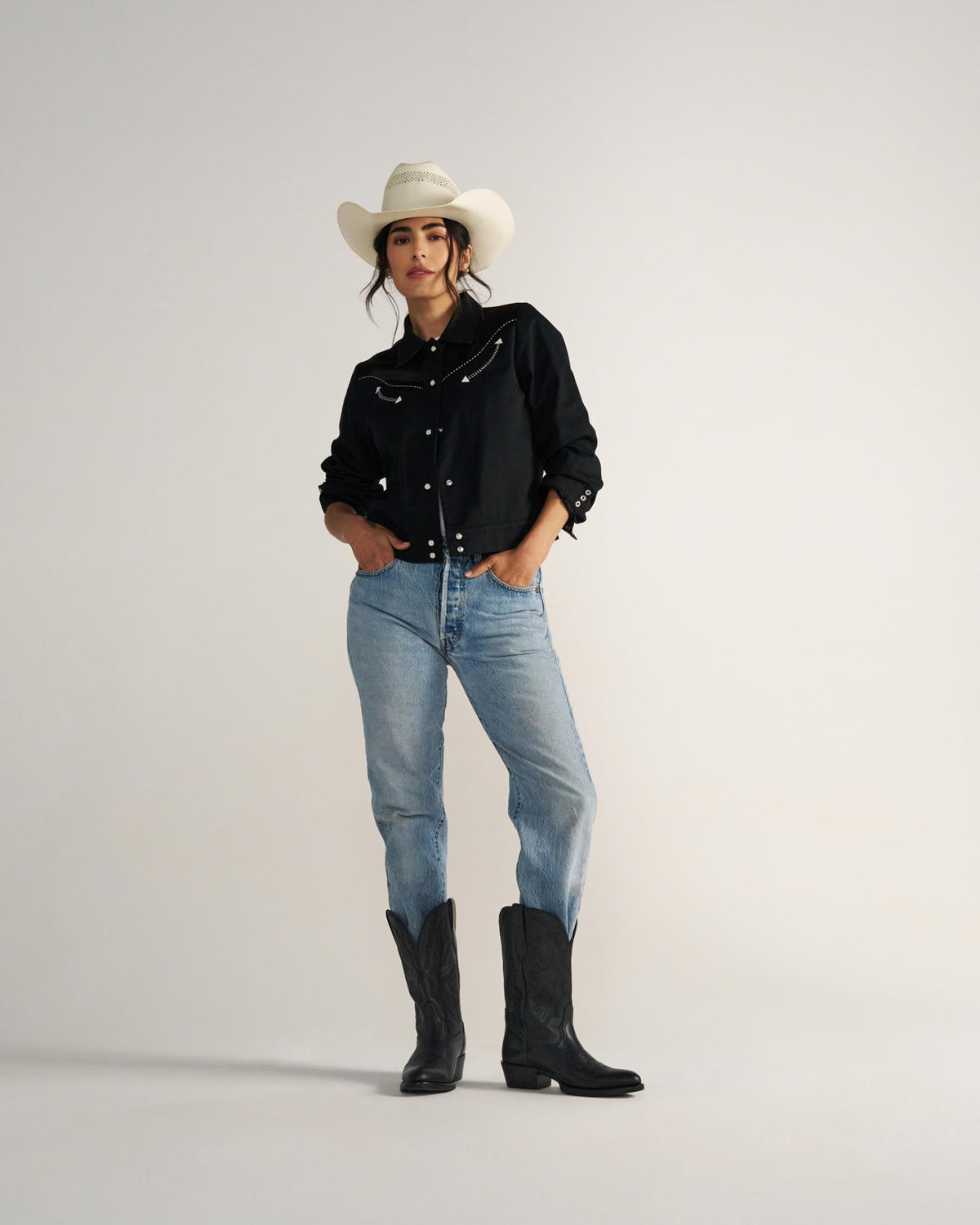 The Britt in Midnight Cowhide. A woman stands against a plain background wearing a white cowboy hat, black shirt, blue jeans, and striking Midnight Cowhide boots.