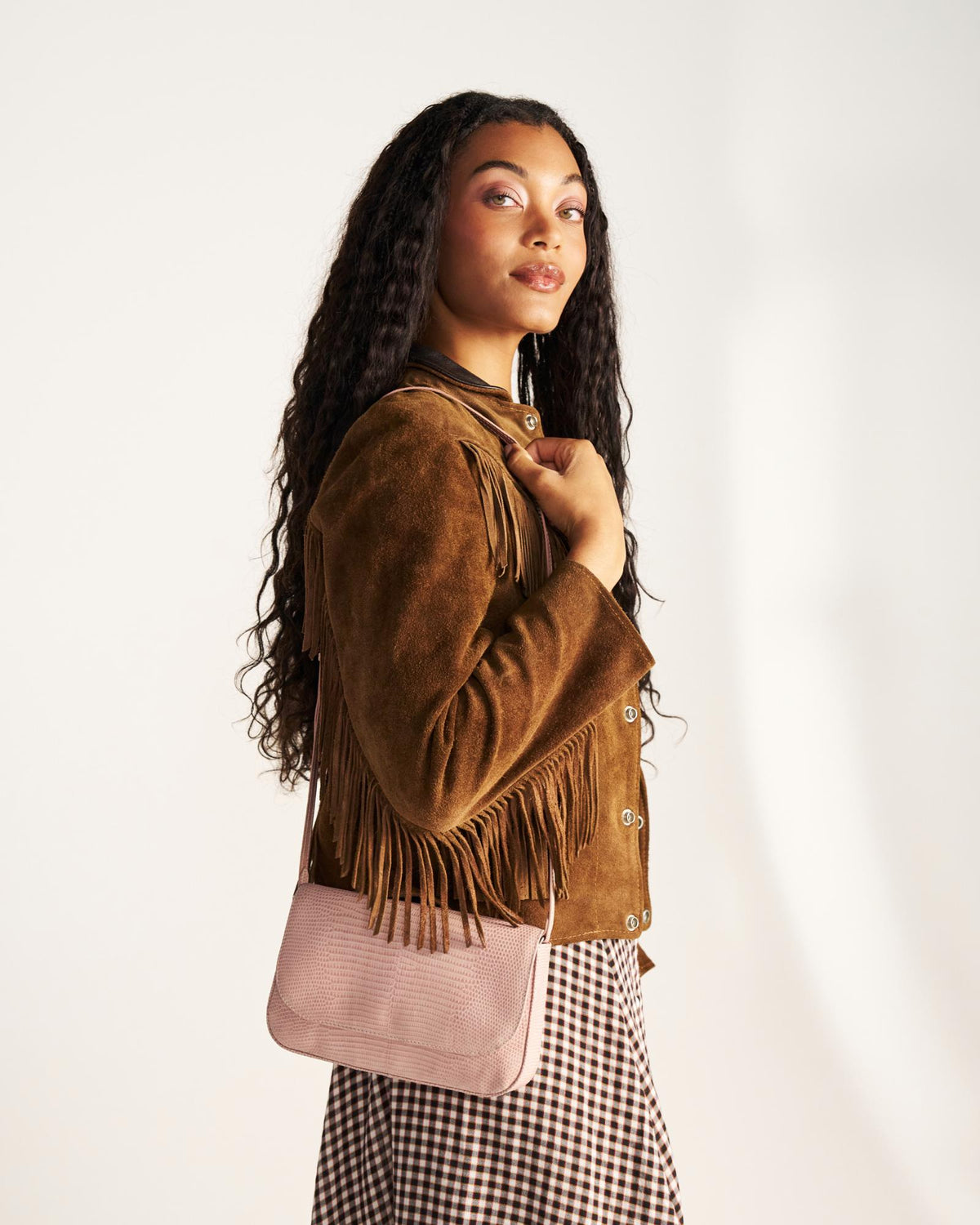 A woman with long curly hair wearing a brown fringed suede jacket and a checkered skirt, holding a light pink shoulder bag, stands against a plain background.