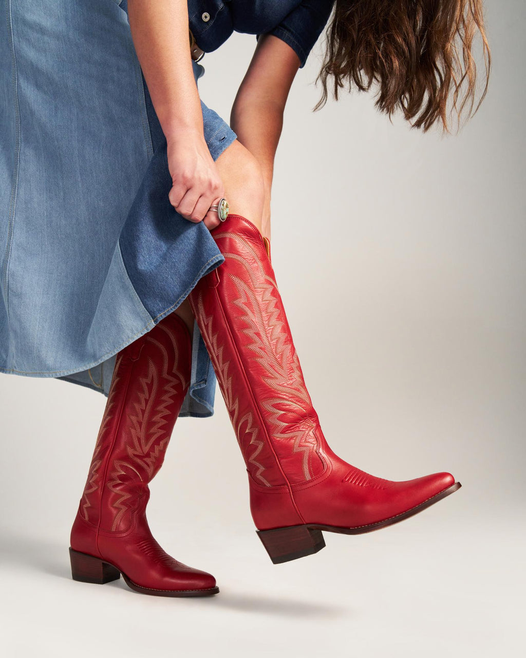 Person wearing a blue denim dress bends down to adjust a knee-high pair of crimson western cowboy boots, The Abby Wide Calf Cowhide, detailed with decorative stitching.