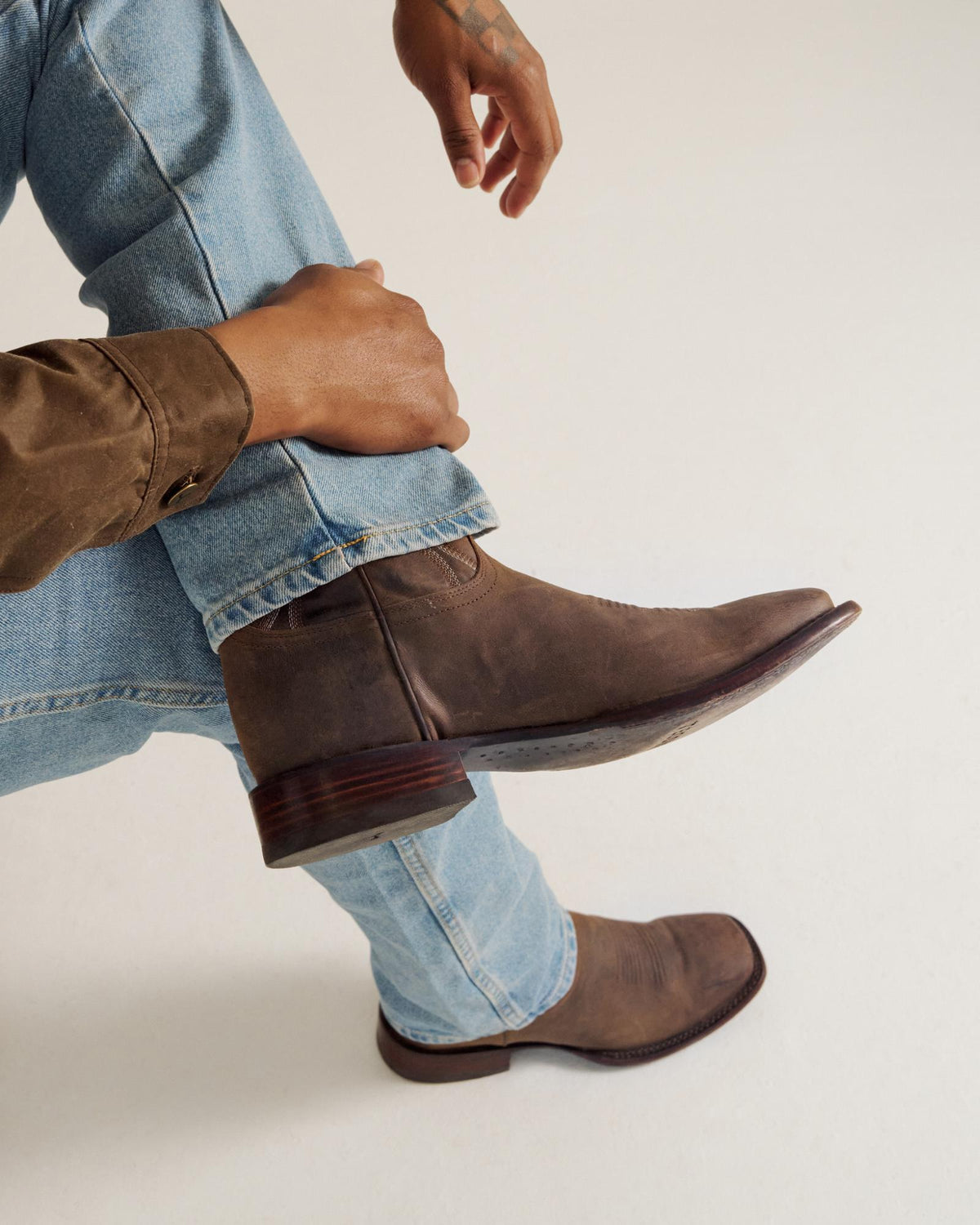 Person wearing light blue jeans, a brown shirt, and brown leather boots, sitting with one leg crossed over the other against a plain light background.