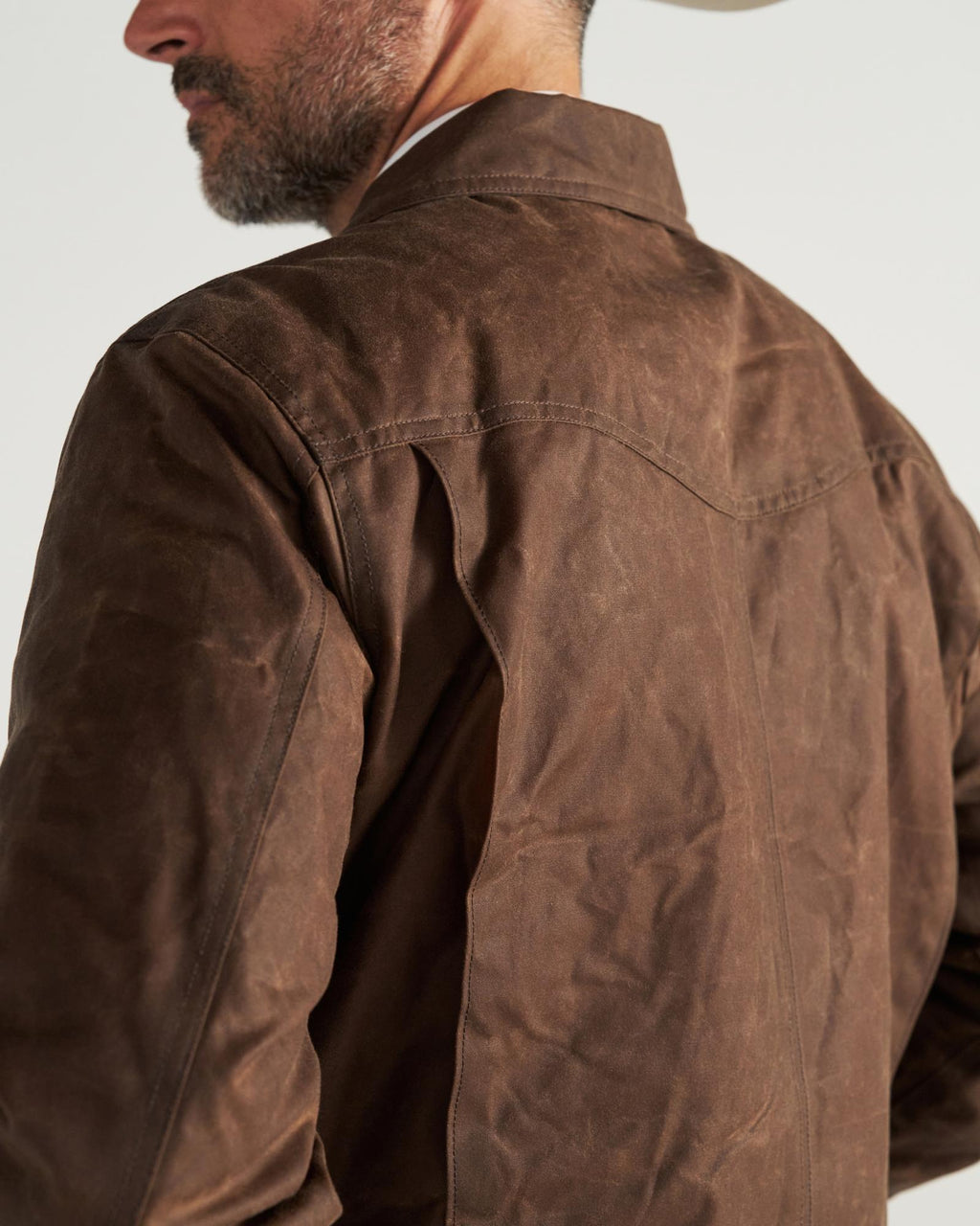A man wearing a brown waxed canvas jacket, viewed from the back and side, with a partial view of his face and a hat.