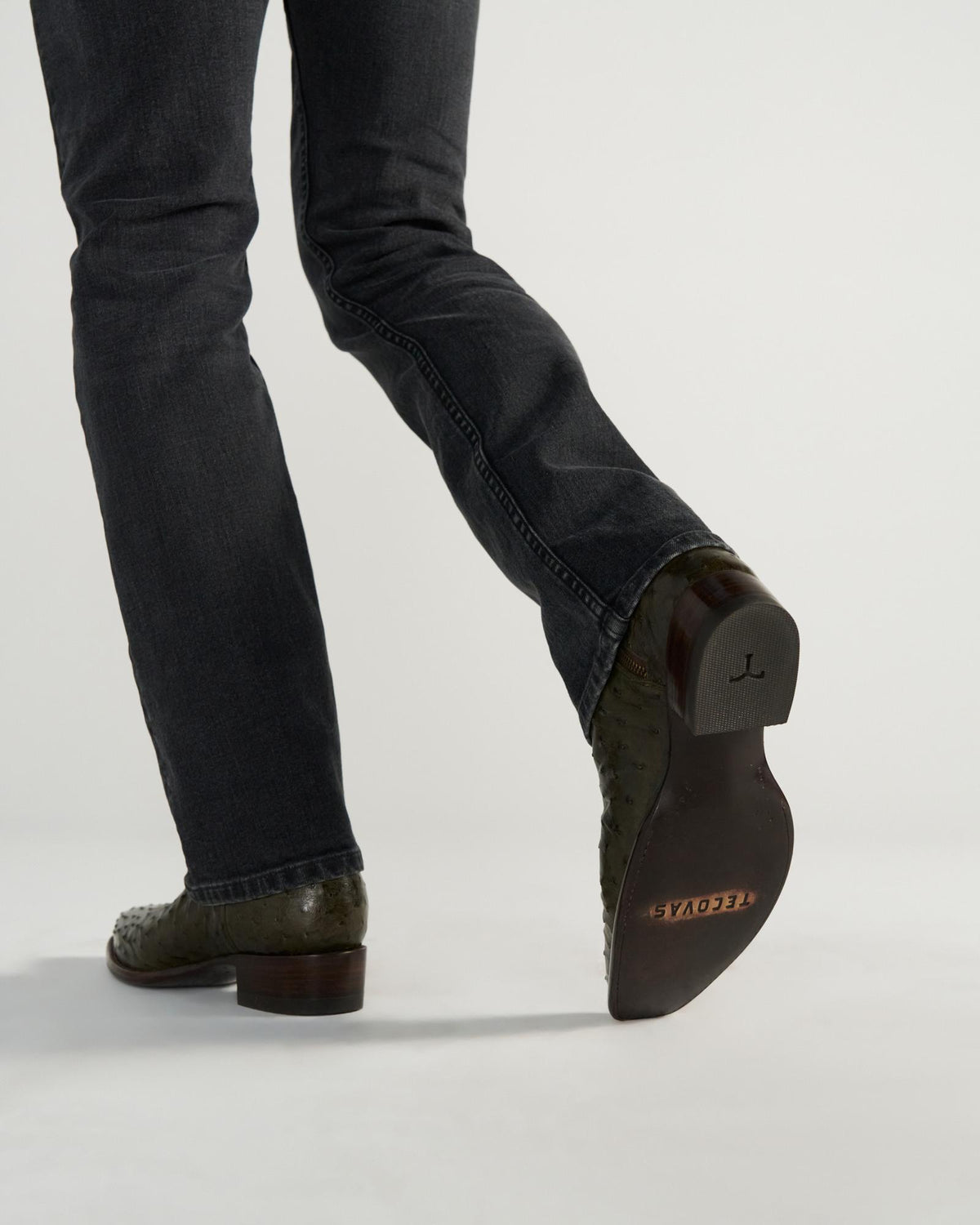 The Jack in Dark Forest Ostrich. Person wearing dark jeans and textured leather boots—crafted from premium Dark Forest Ostrich leather—with one foot lifted to show the sole, which has "TECOYAS" printed on it.