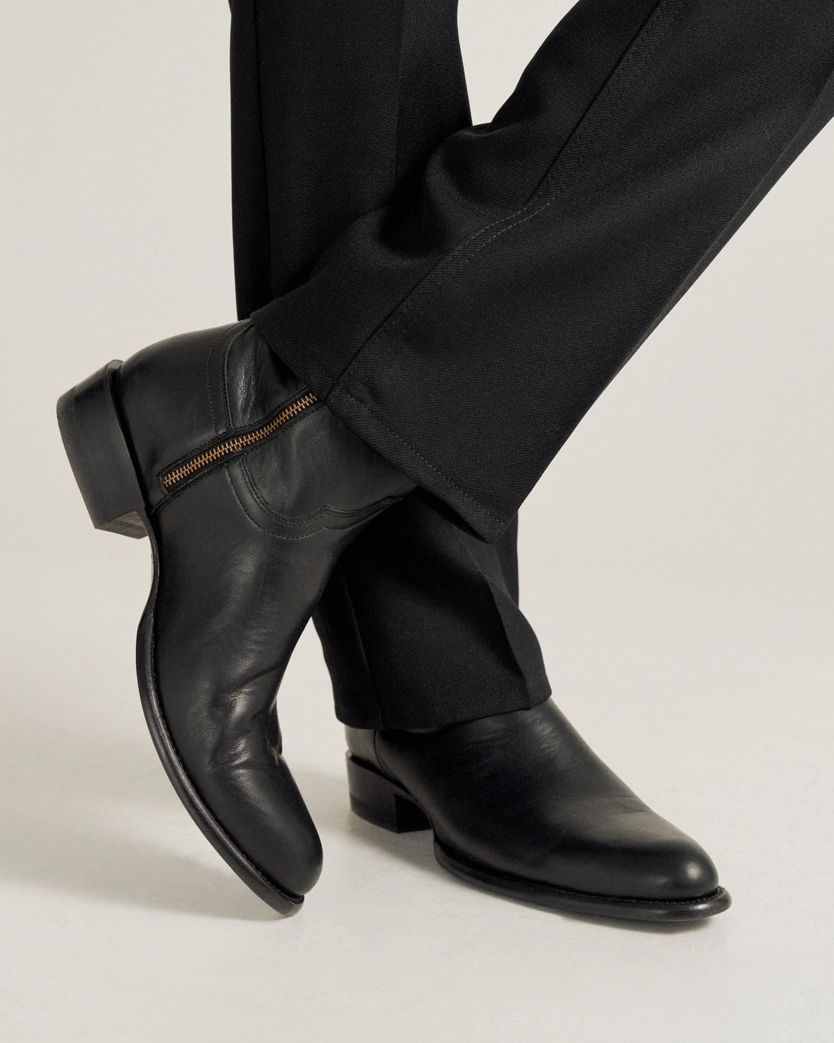 A person wearing black dress pants and black leather boots with a side zipper, standing with one leg crossed over the other.