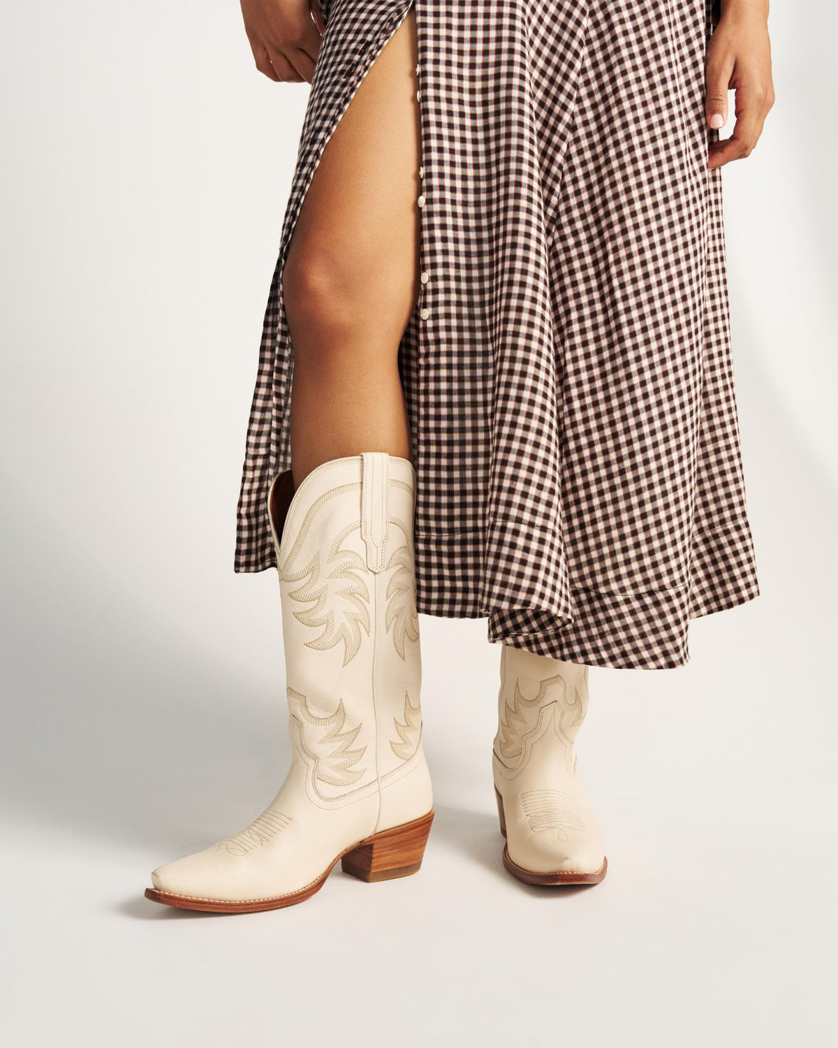 The Annie in Bone Cowhide. Person wearing a brown and white gingham skirt with a high slit and cream-colored bone cowhide cowboy boots, standing against a plain background.
