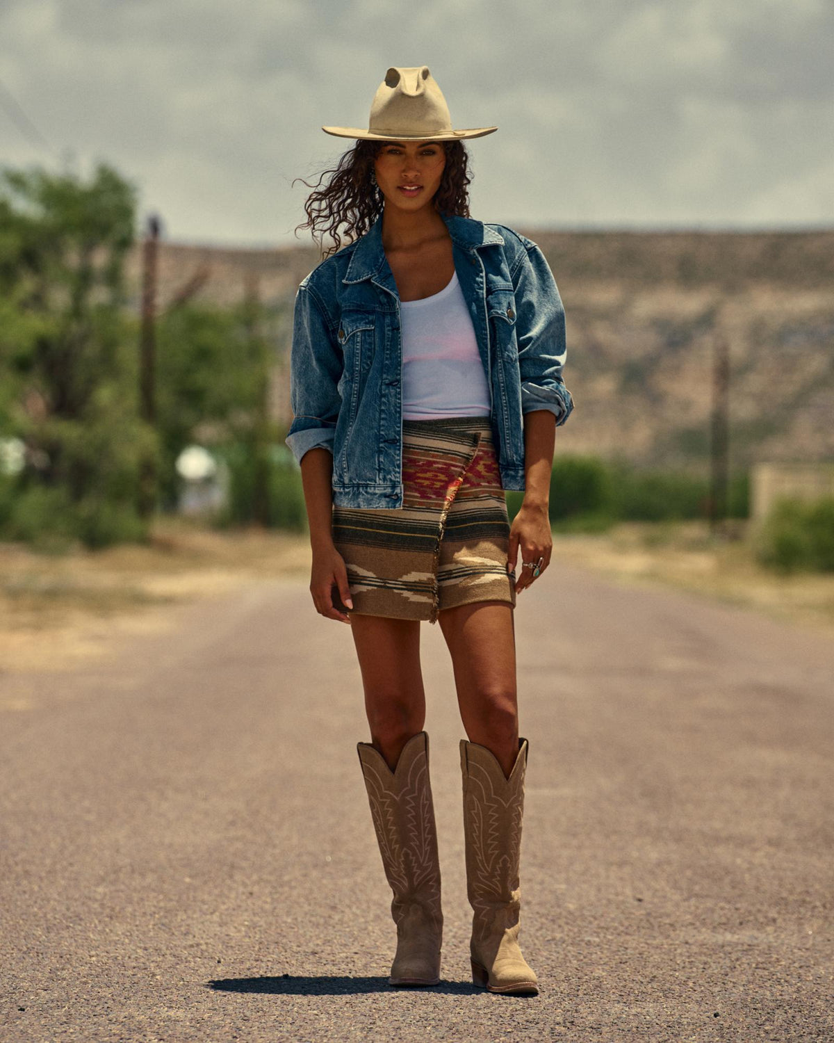 The Abby in Driftwood Suede. A woman stands on a rural road in The Abby Suede cowboy boots, paired with a denim jacket, white tank top, and patterned skirt. Trees and a mountain are visible in the background.