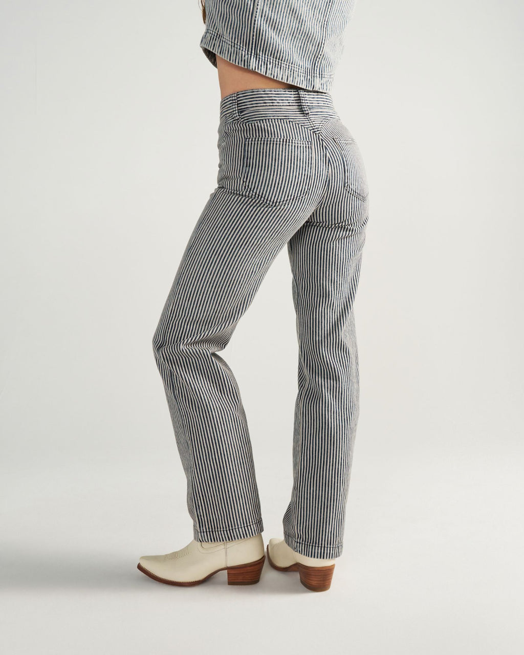 Person wearing striped pants, a matching jacket, and white heeled boots, standing sideways against a plain background.