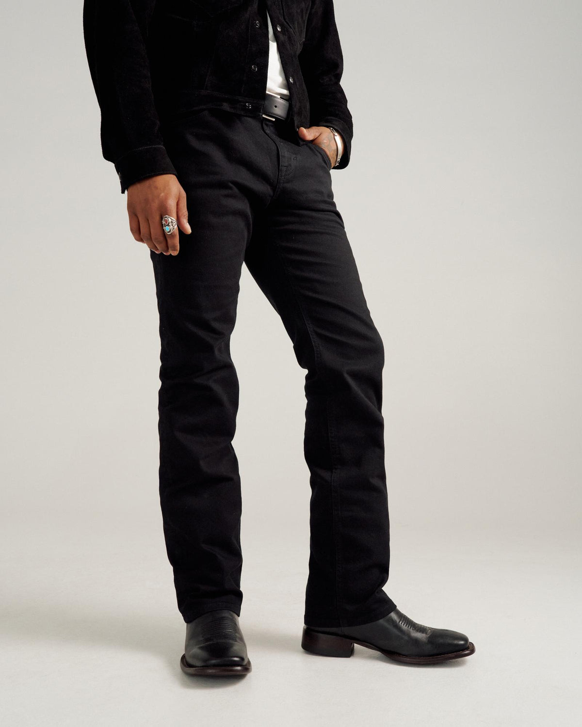 A person stands wearing black jeans, a black jacket, black boots, and a ring, with their hands by their sides against a plain background.