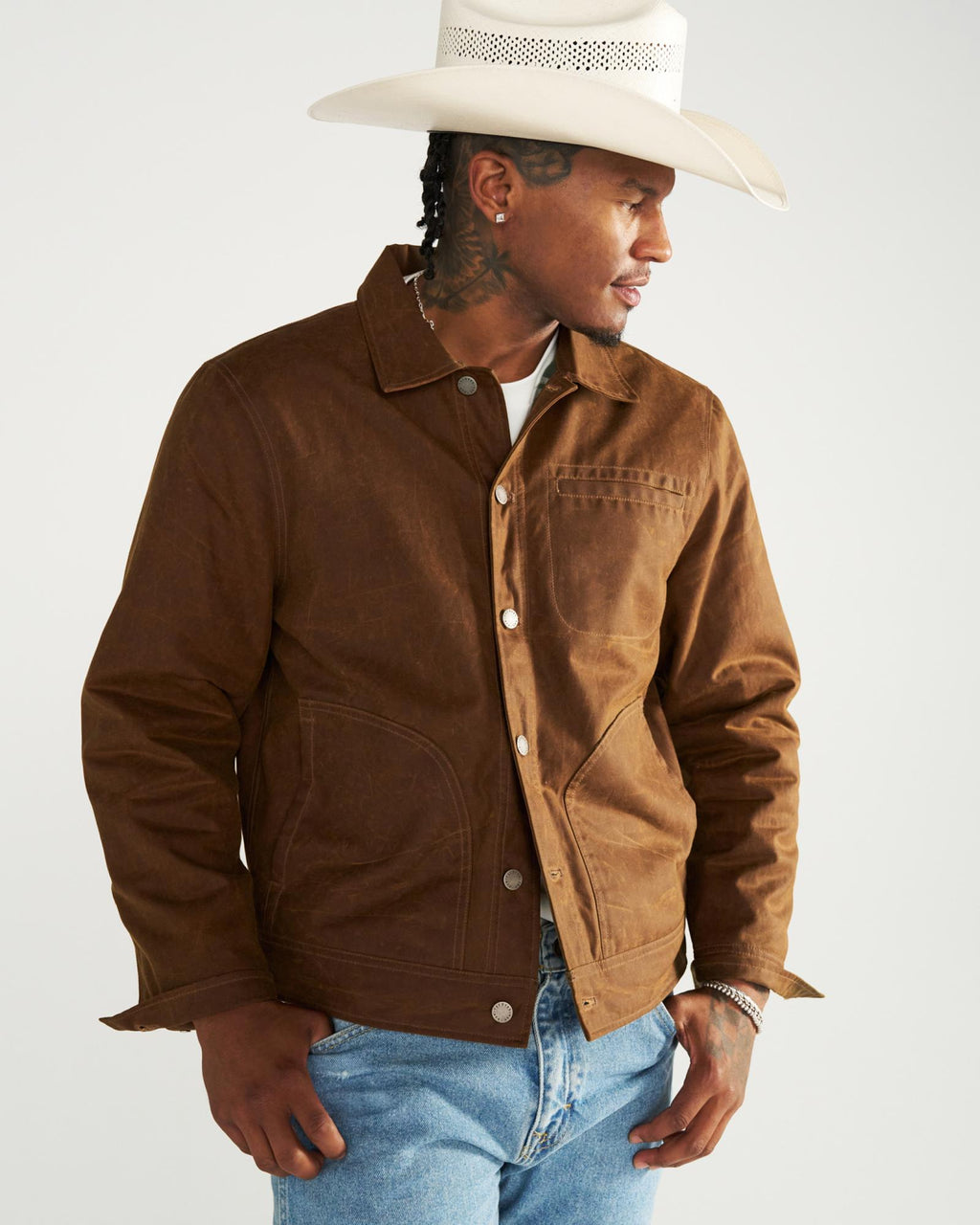 A man wearing a white cowboy hat and a brown canvas chore jacket over a white t-shirt and blue jeans.
