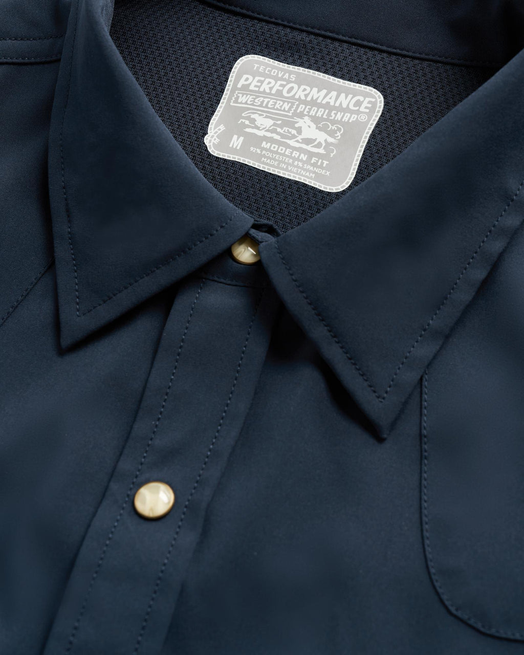 Close-up of a navy blue shirt with gold snap buttons and a white label that reads "Tecovas Performance Western Pearl Snap, Modern Fit, M.