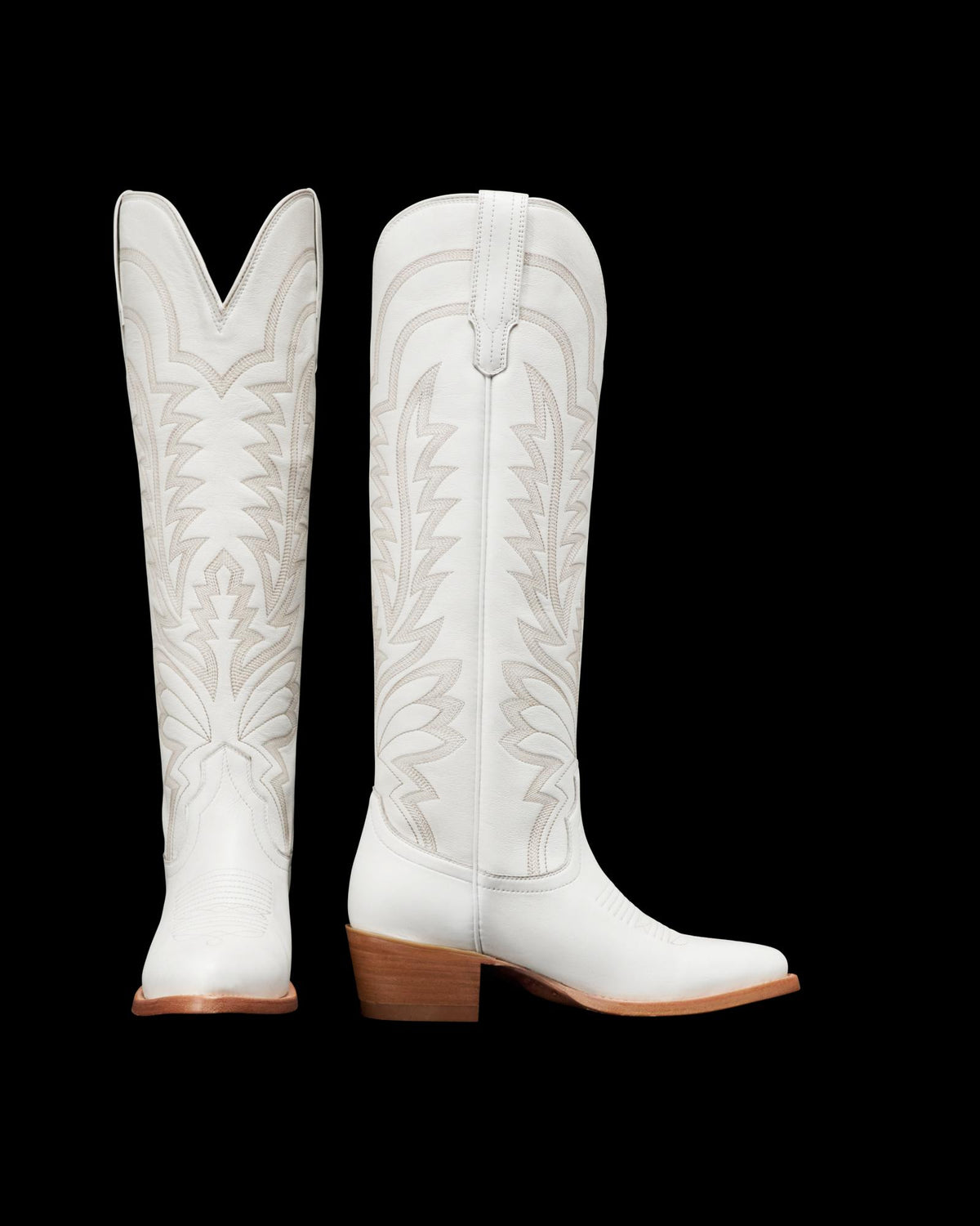 Two white cowboy boots with intricate stitching patterns, wooden heels, and pointed toes. One boot is shown from the back and the other from the side.