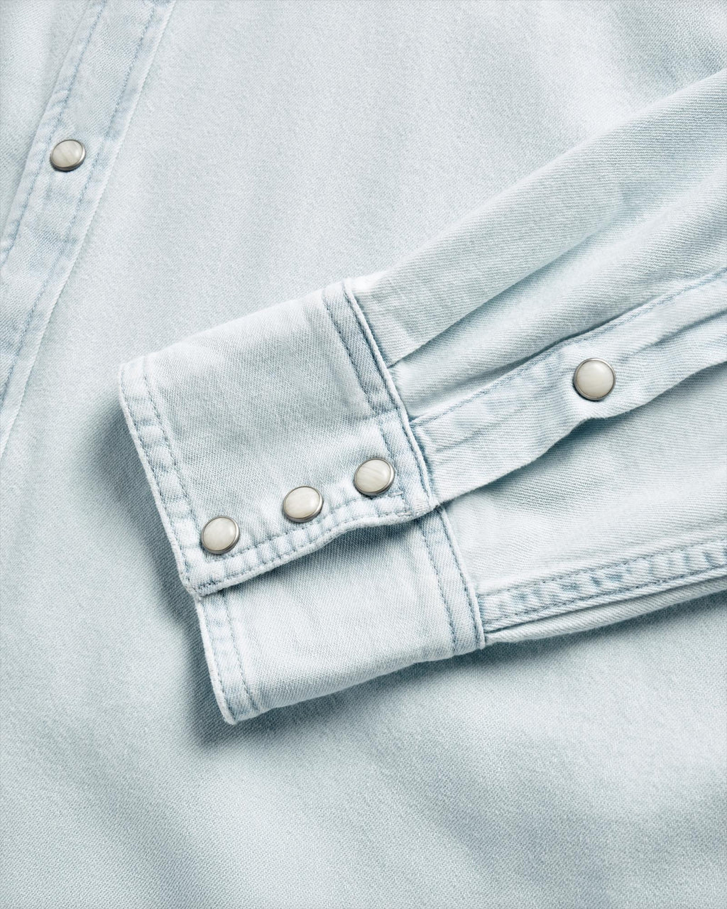 Close-up of a light blue denim shirt sleeve and cuff, featuring metal snap buttons and visible stitching details.