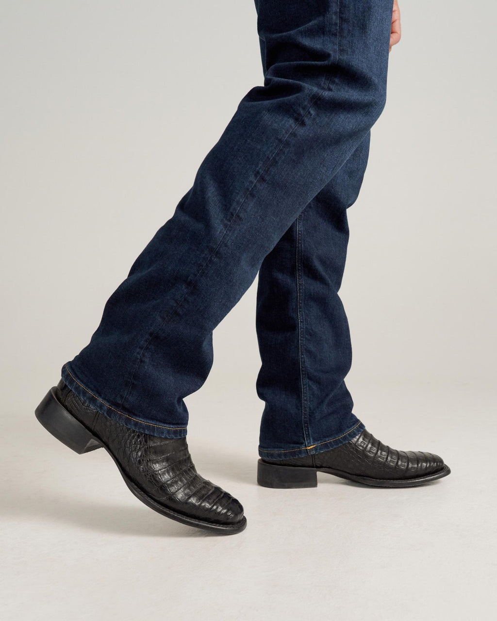 A person wearing midnight blue jeans and The William Caiman black crocodile-patterned dress shoes is walking on a light surface.