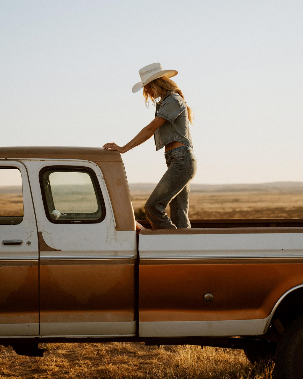 A person wearing a cowboy hat and denim outfit stands in the bed of an old pickup truck parked in a dry, open field.