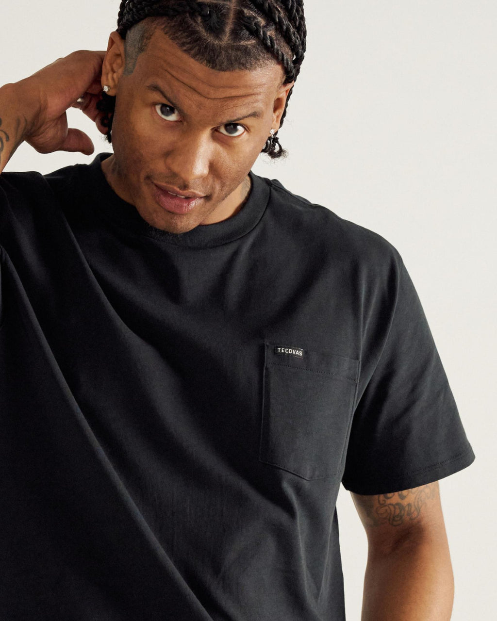 Man with braided hair wearing a black T-shirt with a small chest pocket and "TECOGAN" label, looking at the camera with one hand behind his head.
