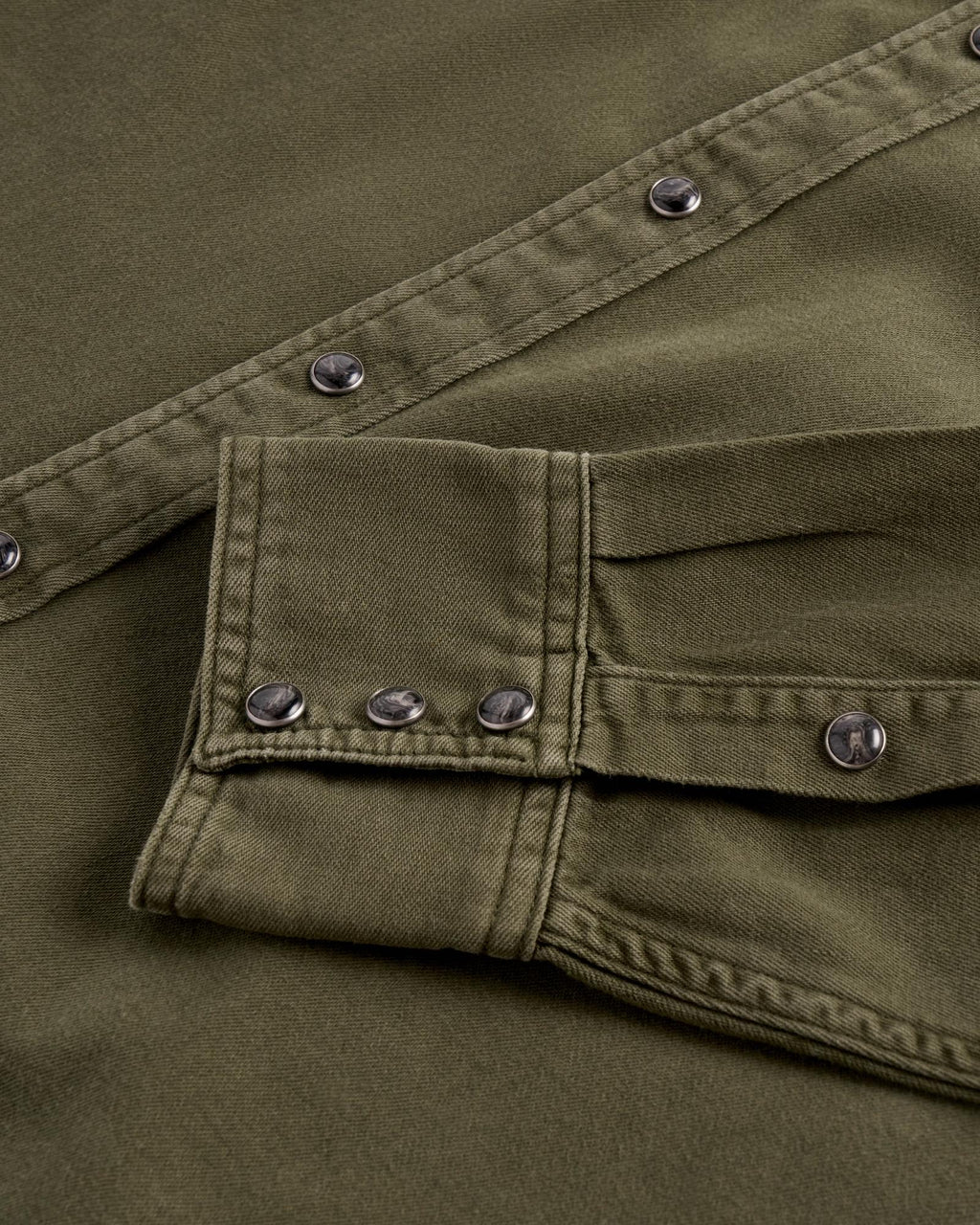 Close-up of an olive green denim shirt with dark, marbled snap buttons on the placket and sleeve cuff.