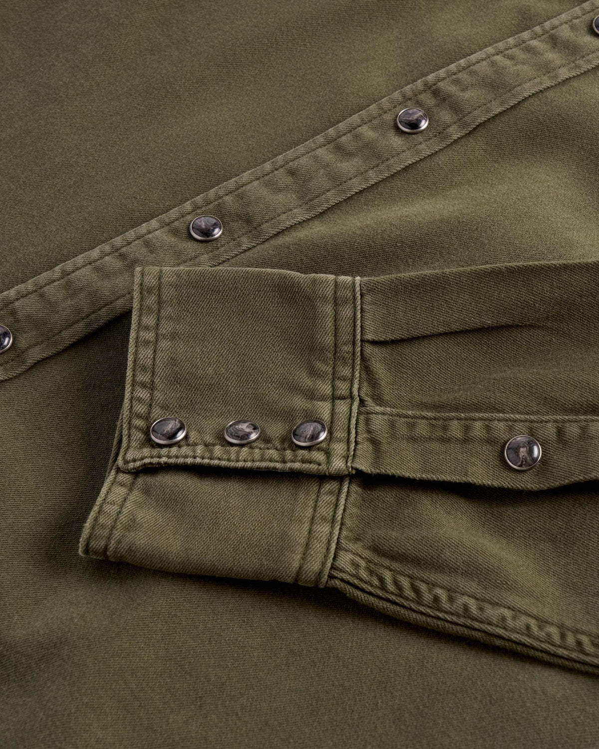 Close-up of an olive green denim shirt with dark, marbled snap buttons on the placket and sleeve cuff.
