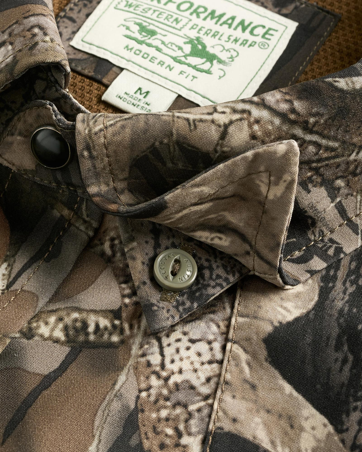 Close-up of a camouflage-patterned shirt with a buttoned collar and visible label reading “PERFORMANCE Western Pearl Snap, Modern Fit, M, Made in Bangladesh.”.