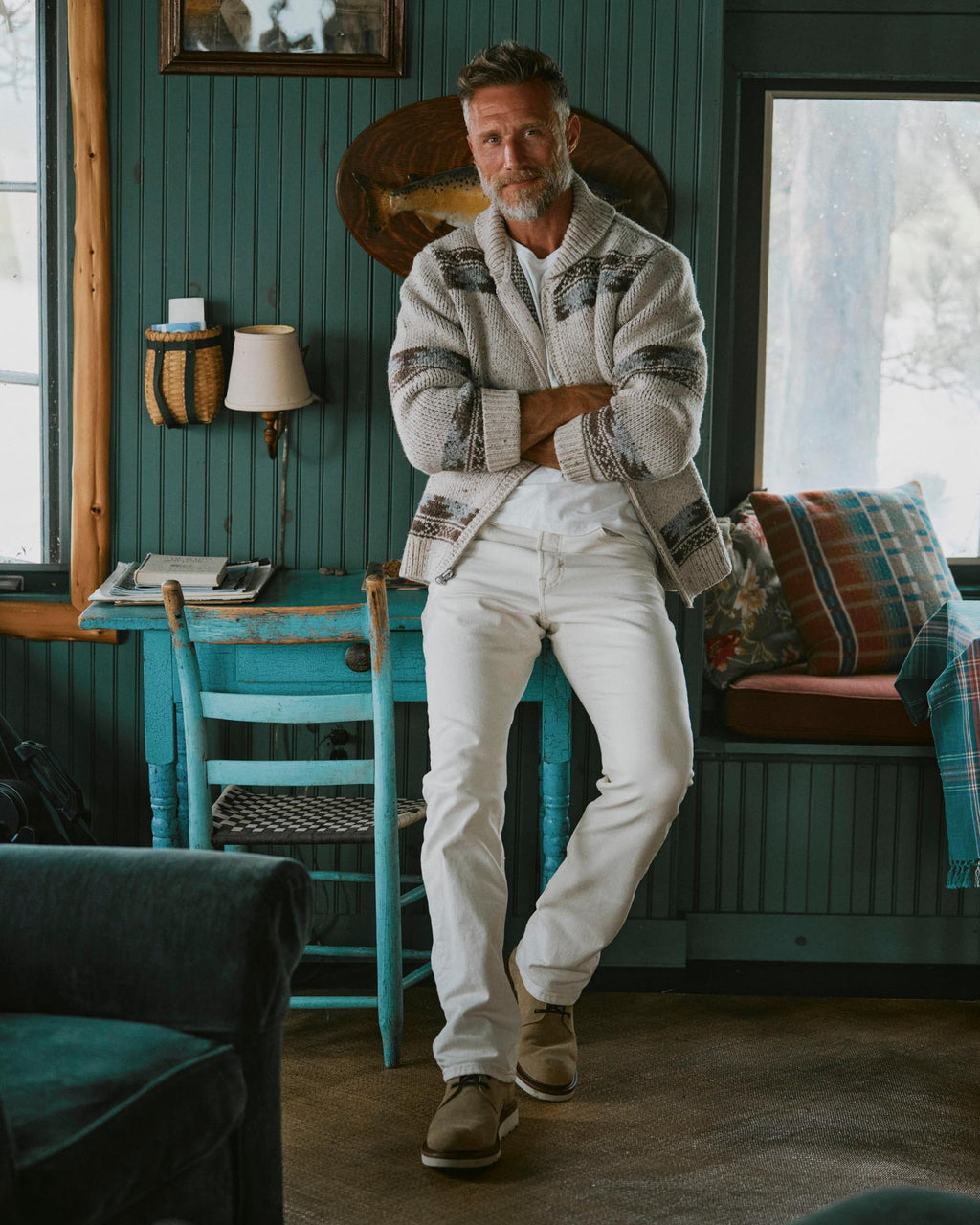 The Knox Lace Up Shearling in Sandstone Roughout. Man with gray hair and beard, wearing a patterned cardigan and white pants, stands casually indoors by a turquoise chair, arms crossed, with windows and sandstone roughout wooden furniture in the background.