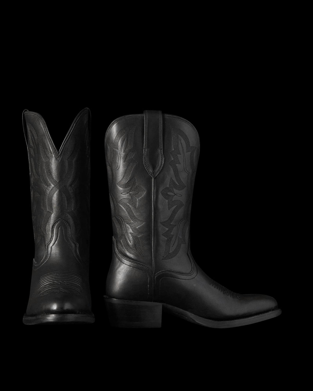 The Britt in Midnight Cowhide. Two black leather cowboy boots crafted from midnight cowhide with decorative stitching are shown; one boot faces forward, while the other is viewed from the side.