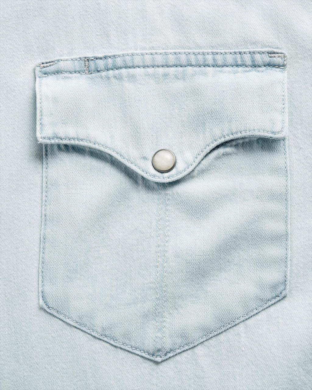 Close-up of a light blue denim shirt pocket with a flap and a metal snap button.