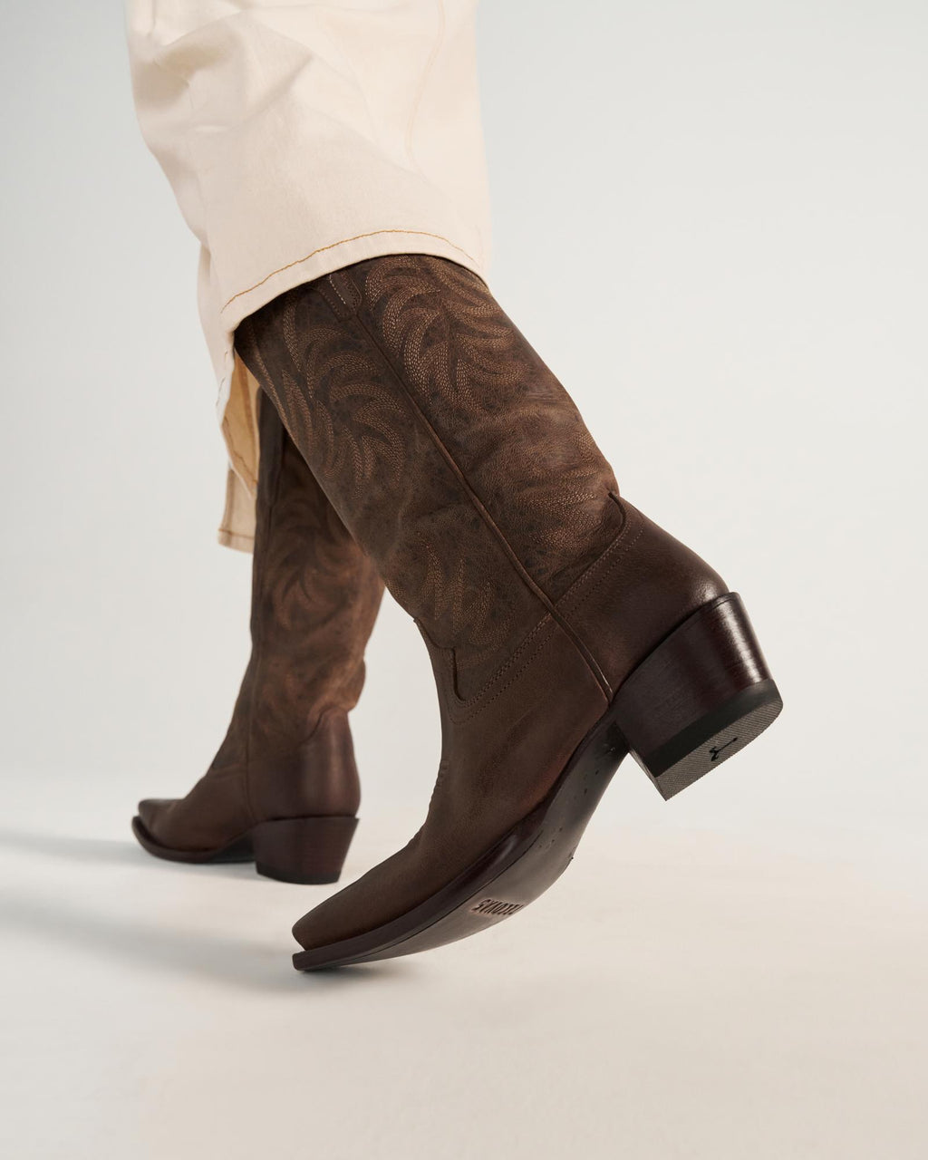 The Annie in Cafe Goat. Person wearing cream-colored pants and brown embroidered leather cowboy boots, photographed from a low angle against a plain background—perfect for a stylish day out at Cafe Goat.